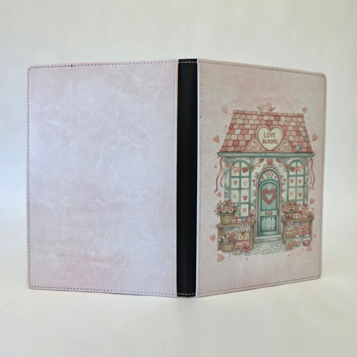Notebook cover with a cute house illustration and 'Love Blooms' text on a heart sign above the door. Pink color theme.