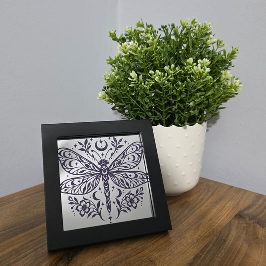 A square black-framed decorative print featuring a purple dragonfly design on a silver reflective background, styled on a wooden table next to a potted green plant against a gray wall.