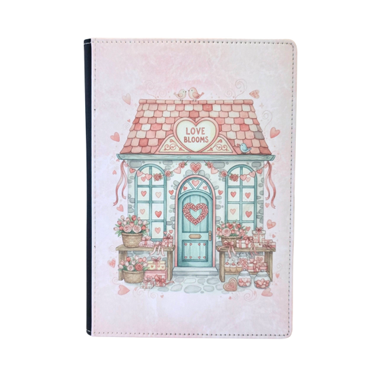 Notebook cover with a cute house illustration and 'Love Blooms' text on a heart sign above the door. Pink color theme.