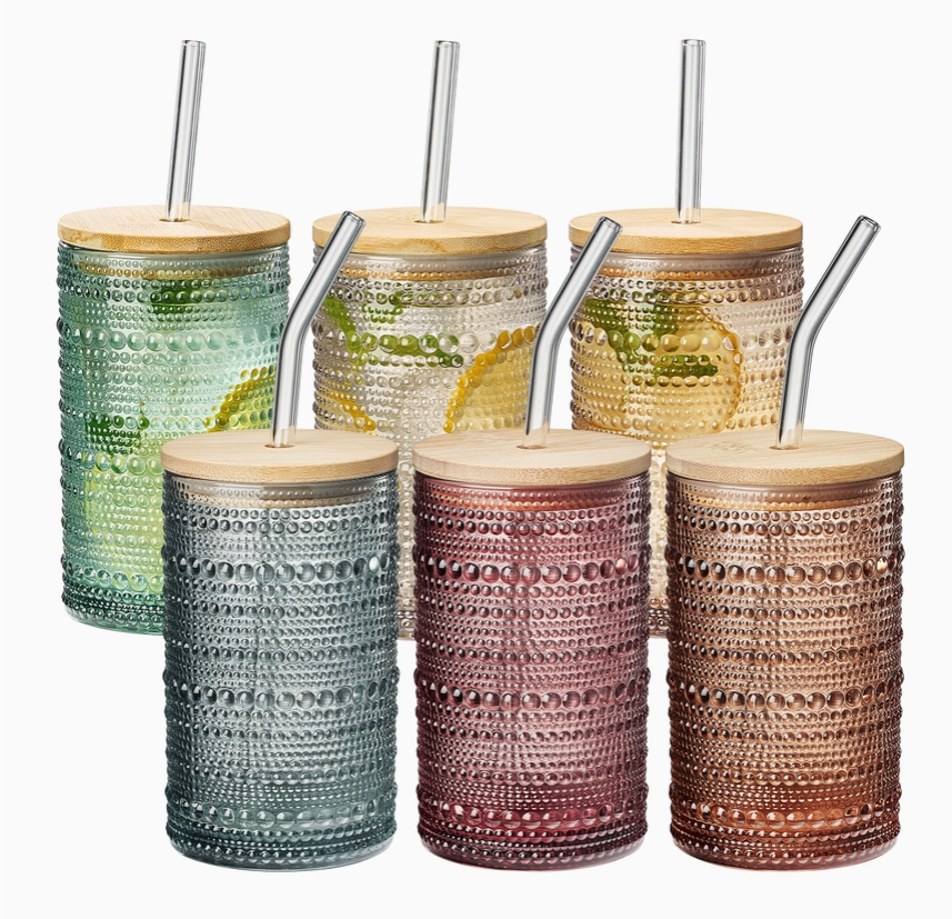 Six patterned glass tumblers with wooden lids and glass straws on a white background. Colors include Green, clear, light pink, blue, reddish purple, and orange.
